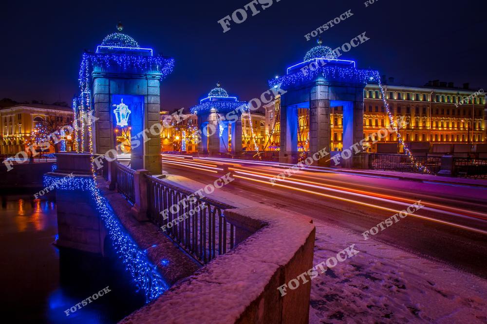 На фотографии изображен мост Ломоносова в Санкт-Петербурге, украшенный праздничной иллюминацией. Мост через Фонтанку особенно красив в зимний период, когда гирлянды и огни подчеркивают его архитектурную изящность. Вечерняя подсветка добавляет ощущение волшебства, а снежный покров усиливает атмосферу зимнего праздника. Это место становится особенно популярным в новогодние и рождественские дни благодаря своей романтичной и уютной обстановке.